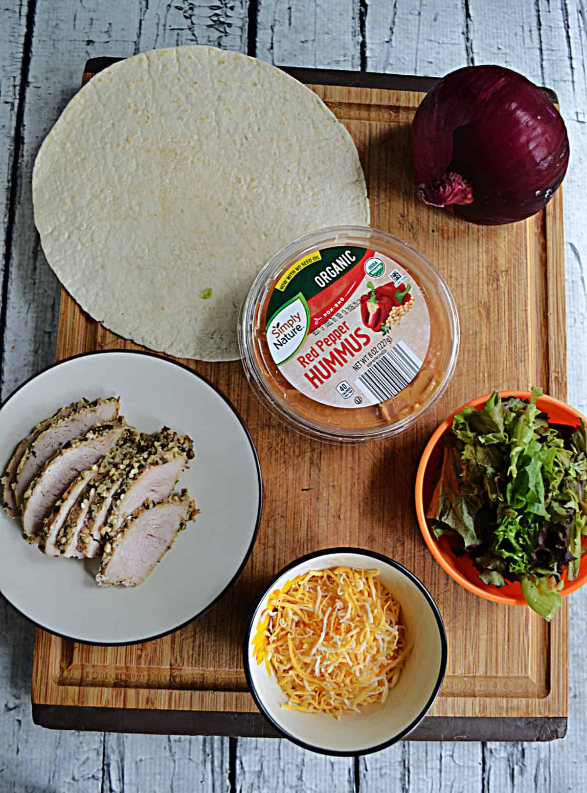 A cutting board with tortillas, sliced turkey, hummus, lettuce, a red onion, and chees eon it.