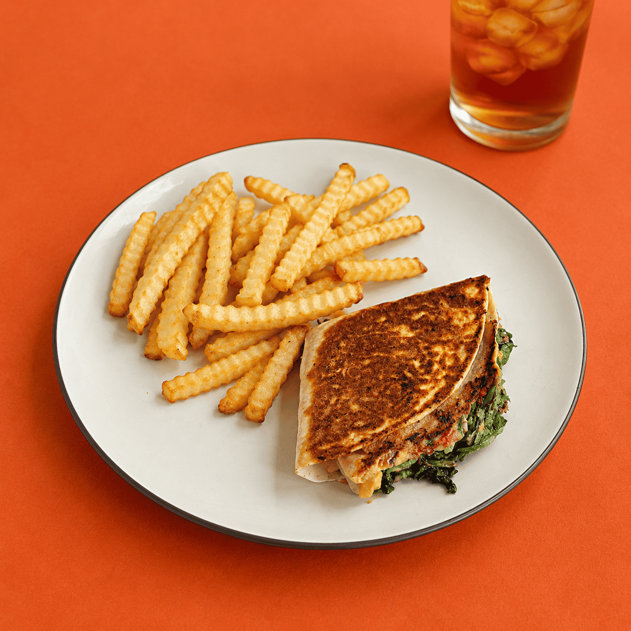 A plate with a golden brown wrap on it along with fries and a glass of tea behind the glass.